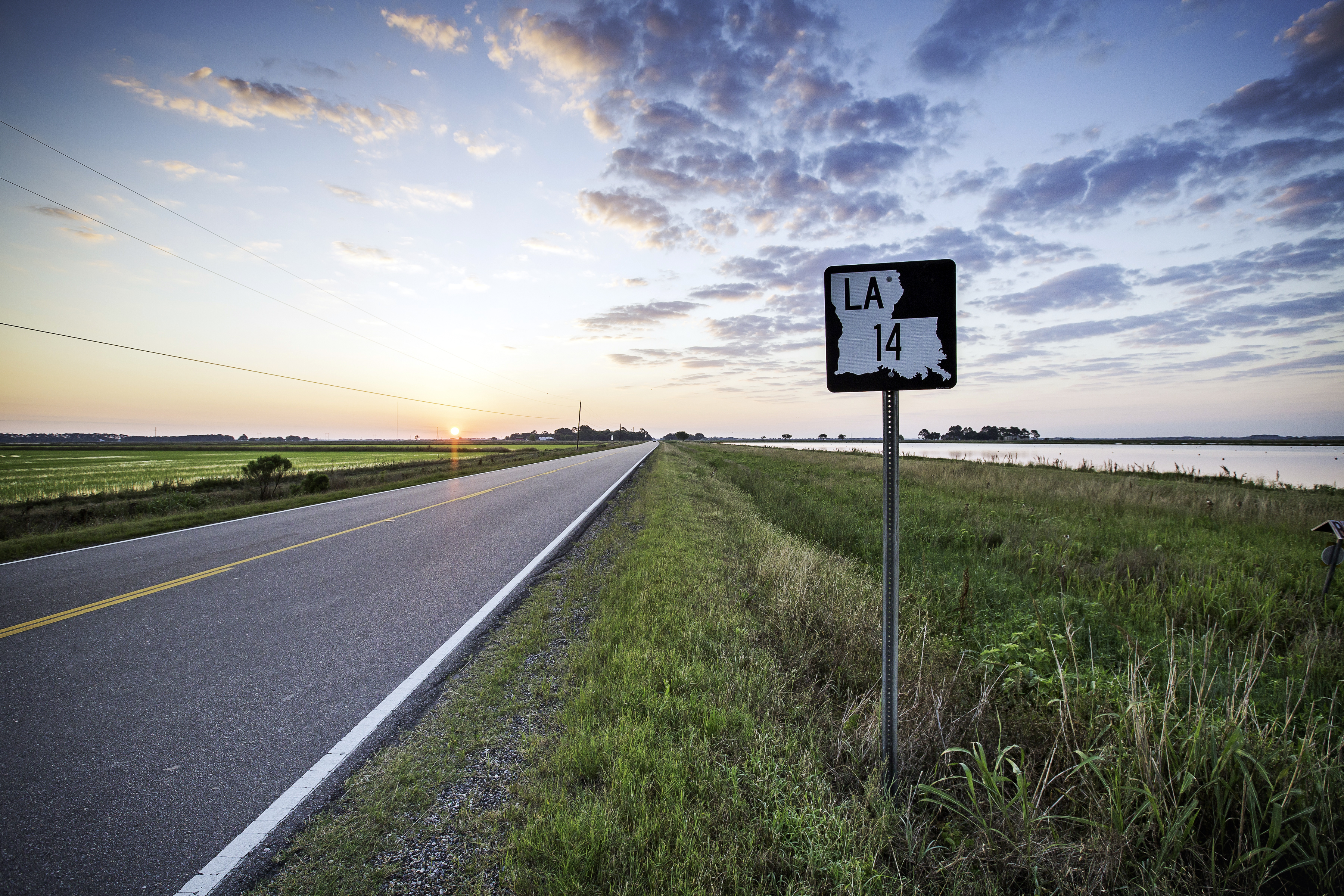 A rural two-lane highway, part of Louisiana's Cajun Corridor Byway, stretches into the distance. In the background the sun peeks over the horizon, brightening a cloudy sky.