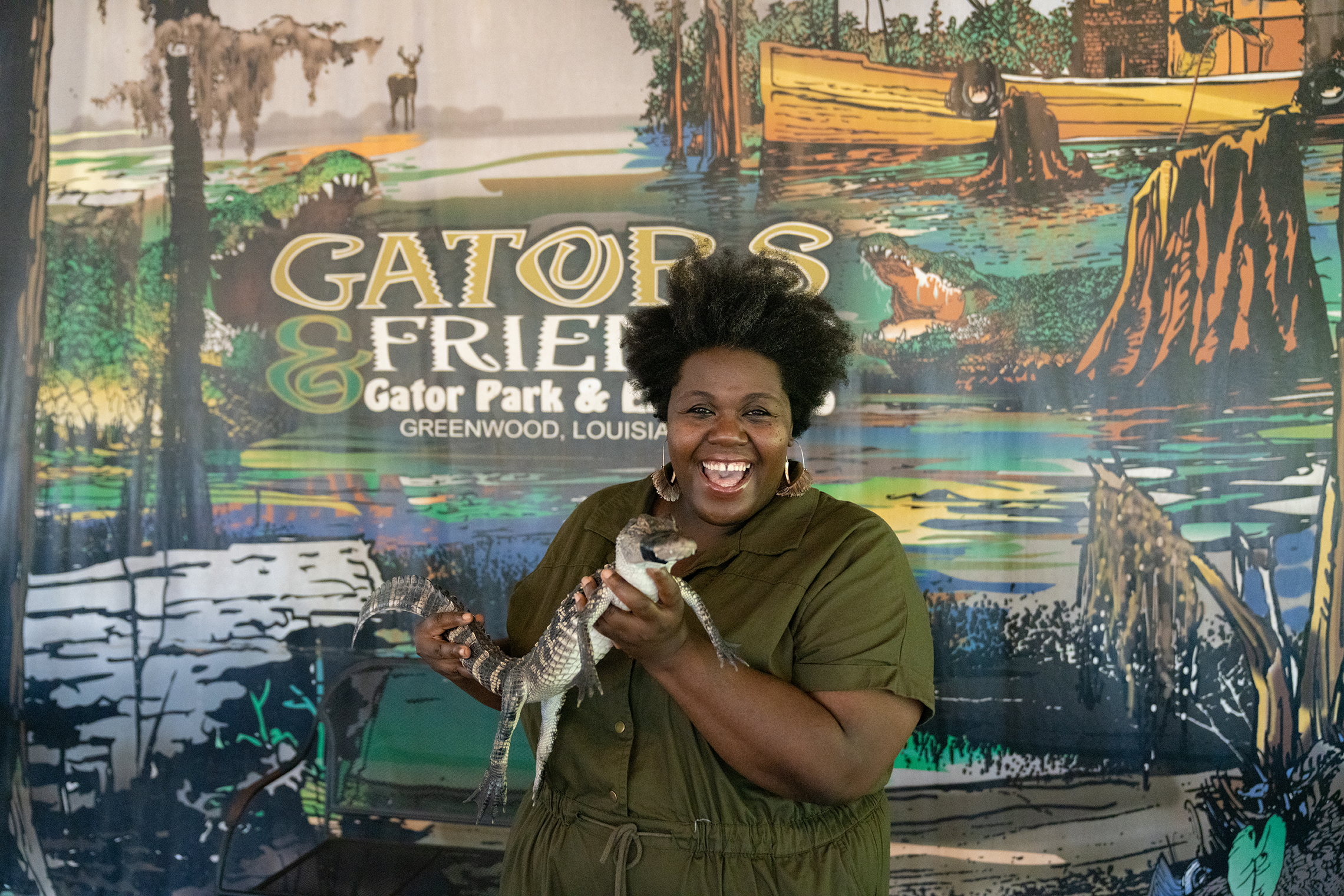 A woman in a green shirt holds a baby alligator in front of a sign advertising Gators & Friends Adventure Park.