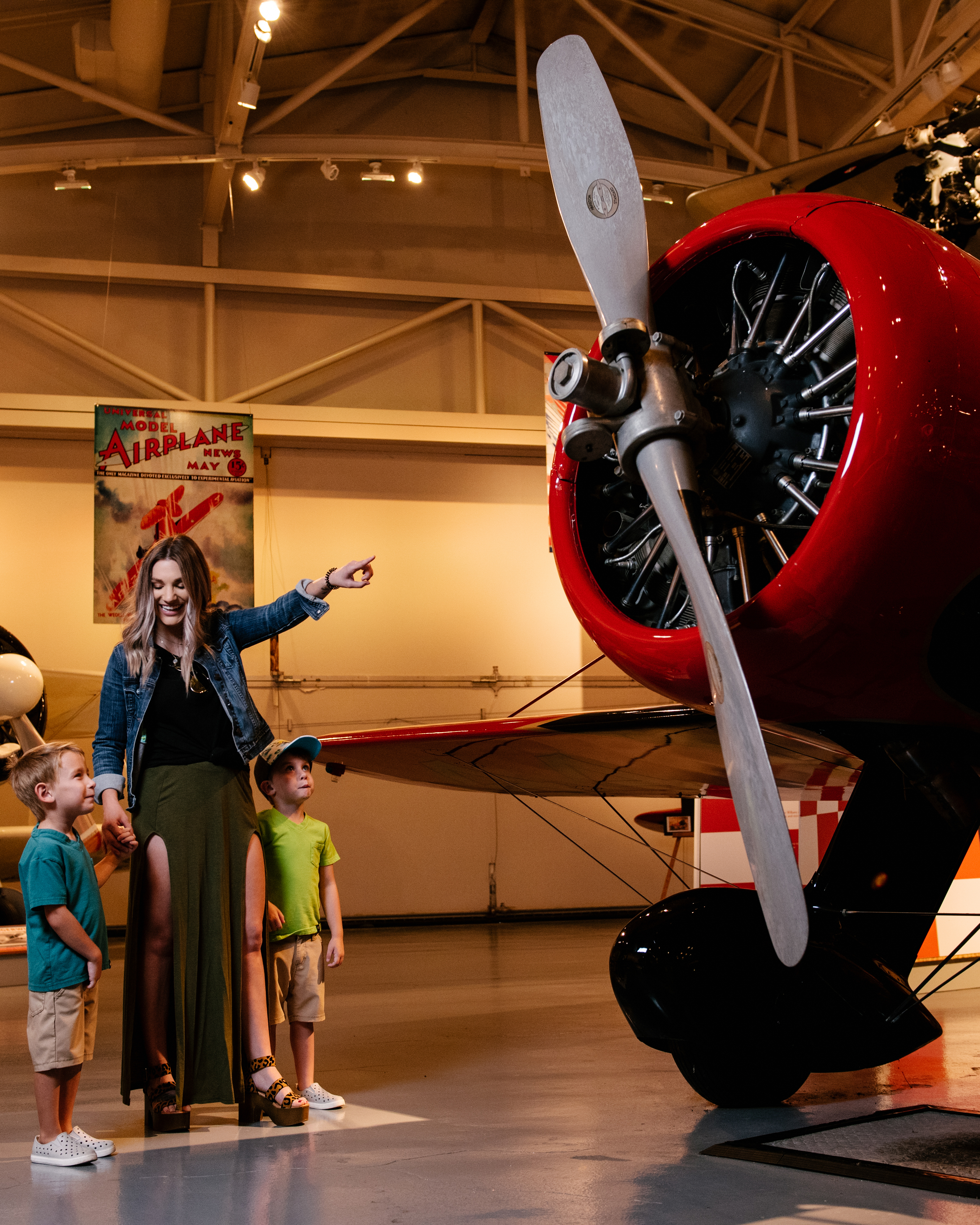 A woman with two children pointing at a large red airplane and its propeller inside a museum