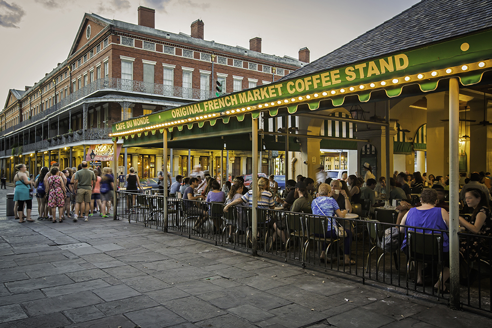 Outdoor view of the lively Cafe du Monde in New Orleans, Louisiana with people sitting at tables under a green canopy.