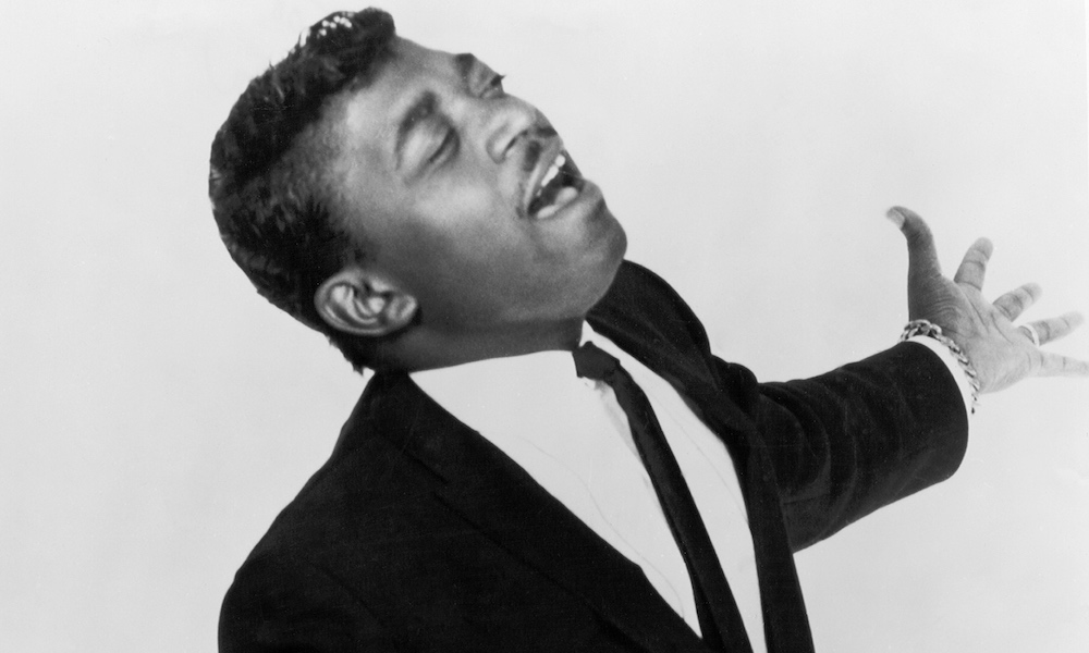 Percy, in a black suit and white shirt, eyes closed, belts out a tune with his arms in the air