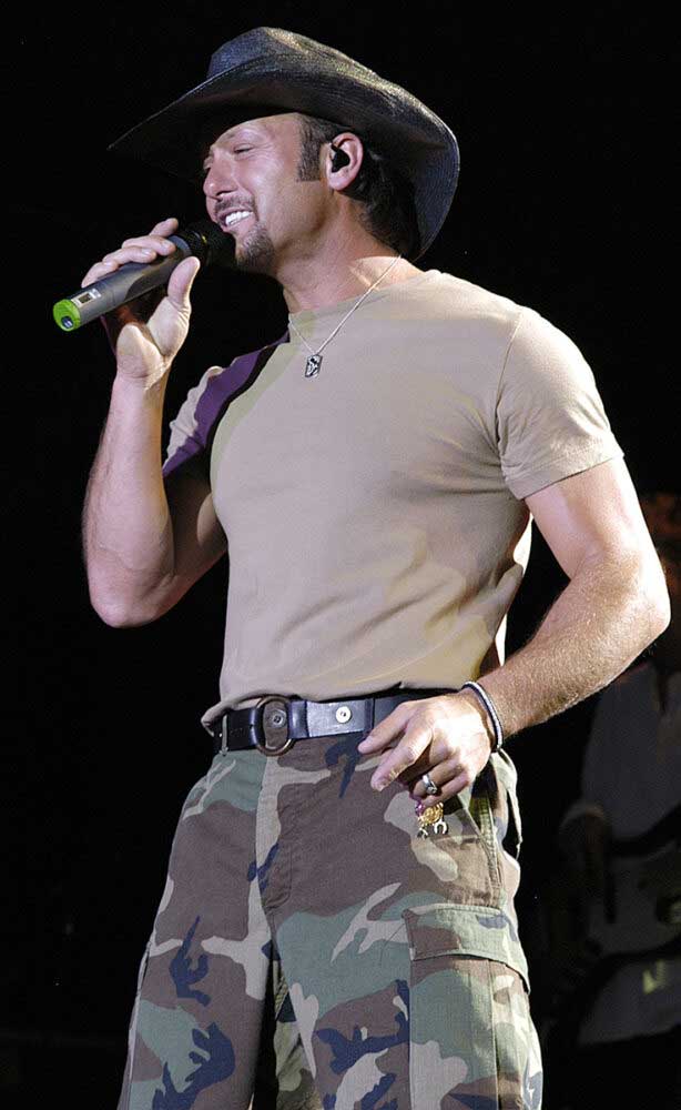 In camp pants, a beige T-shirt and signature black cowboy hat, Tim McGraw holds a mic up to his face while he sings down to a crowd