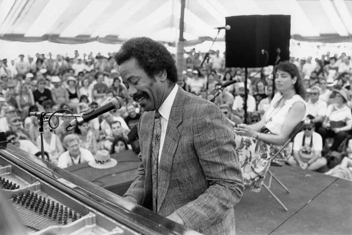 A black-and-white image of Allan playing the piano in front of a crowd gathered under an event tent