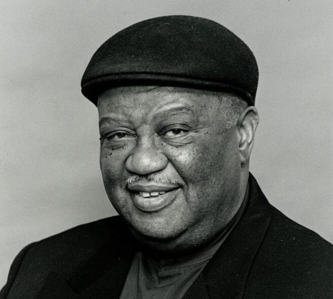 Portrait of Alvin Batiste wearing a dark hat and a jacket with a jaunty kerchief 