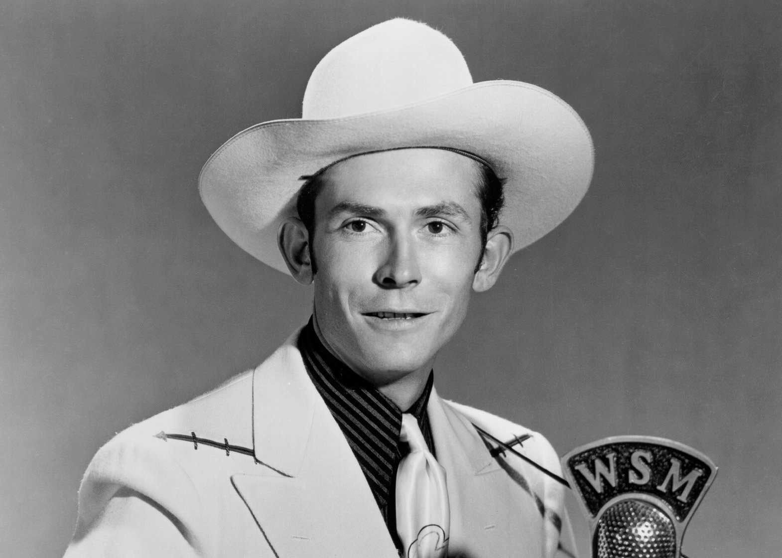 Hank, in a white hat and white suit, plays guitar in front of a mic that says "WSM" on it