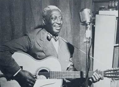 Leadbelly sits with his guitar in front of a microphone in a recording studio