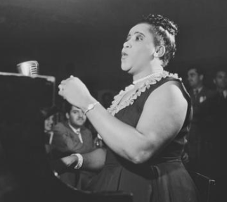 Nellie, in pearls and a black dress, hair in a bun, sings before a gathered audience