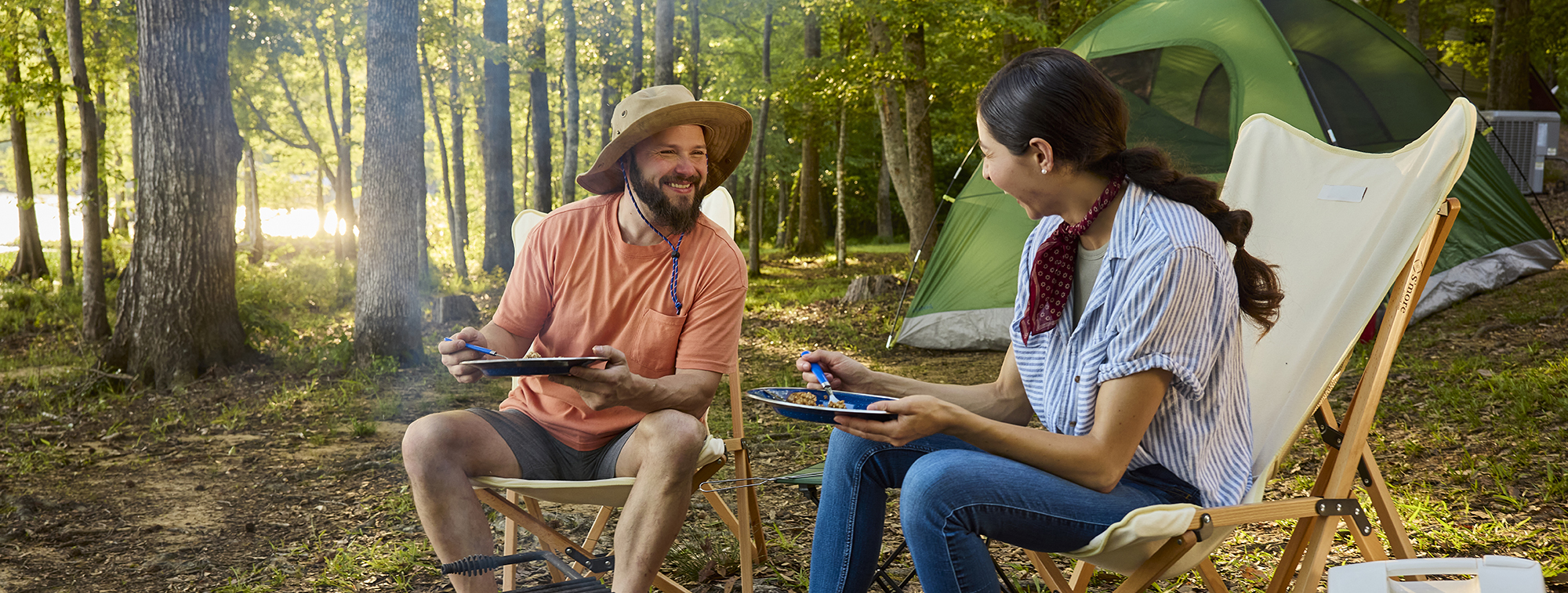 A couple sitting fire side at a camp site with tent in the background enjoying dinner at sunset