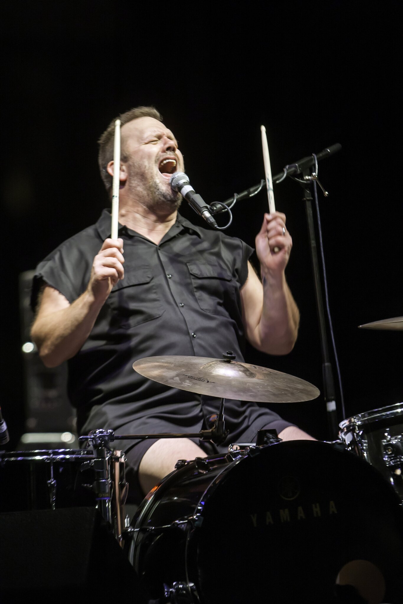 A drummer in a black collared shirt raises his drumsticks while singing at the Red River Revel Arts Festival.