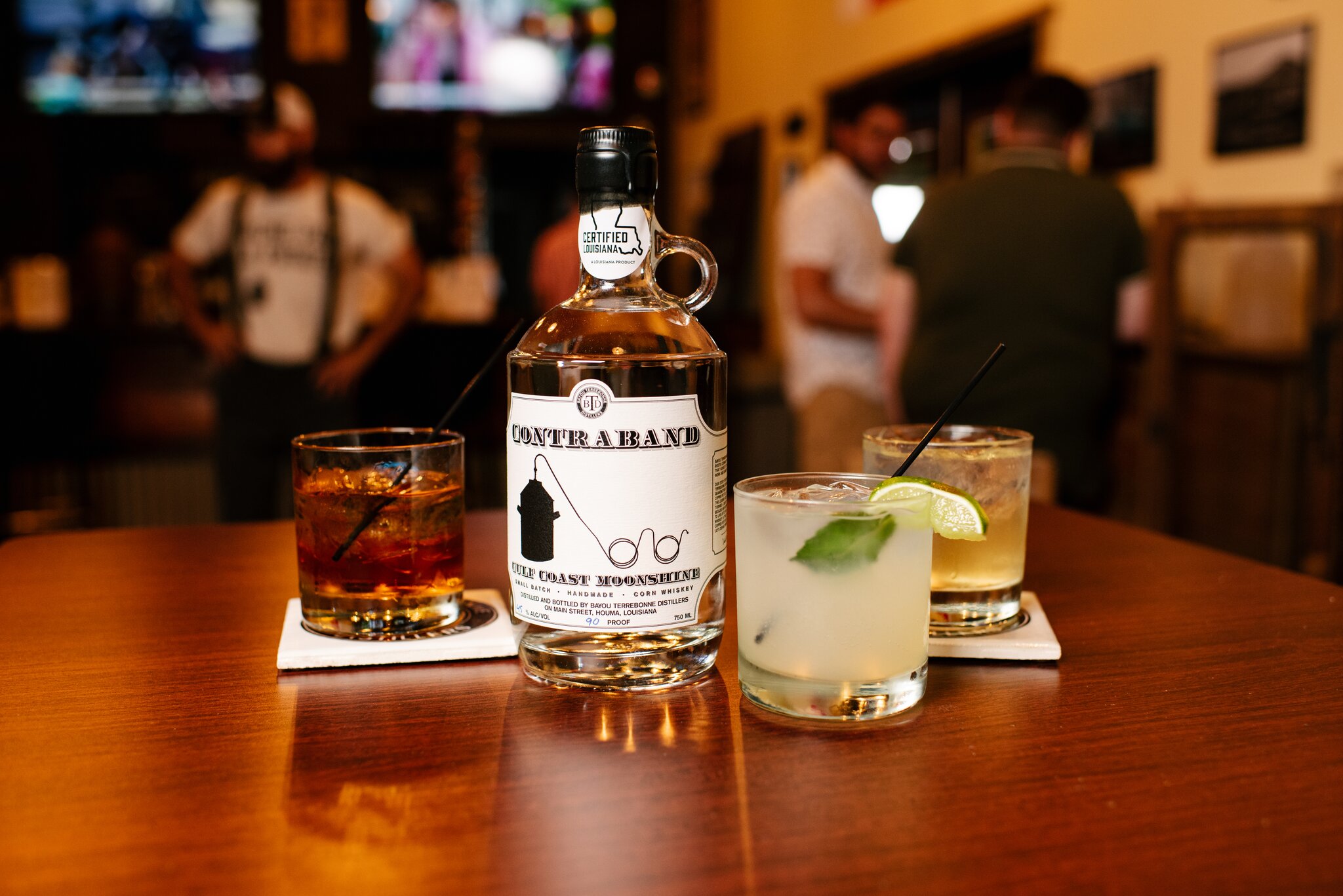 A bottle of moonshine sits next to a variety of cocktails on a wooden table at Bayou Terrebonne Distillers.