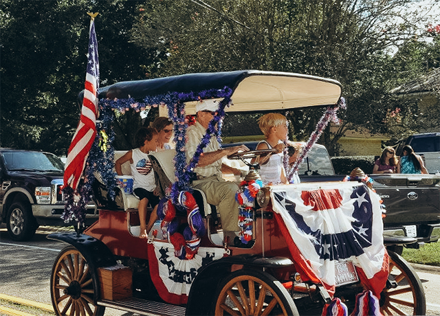 A family drives in an old-fashioned car fully decorated for the Fourth of July, with an American flag proudly displayed at the back of the vehicle.