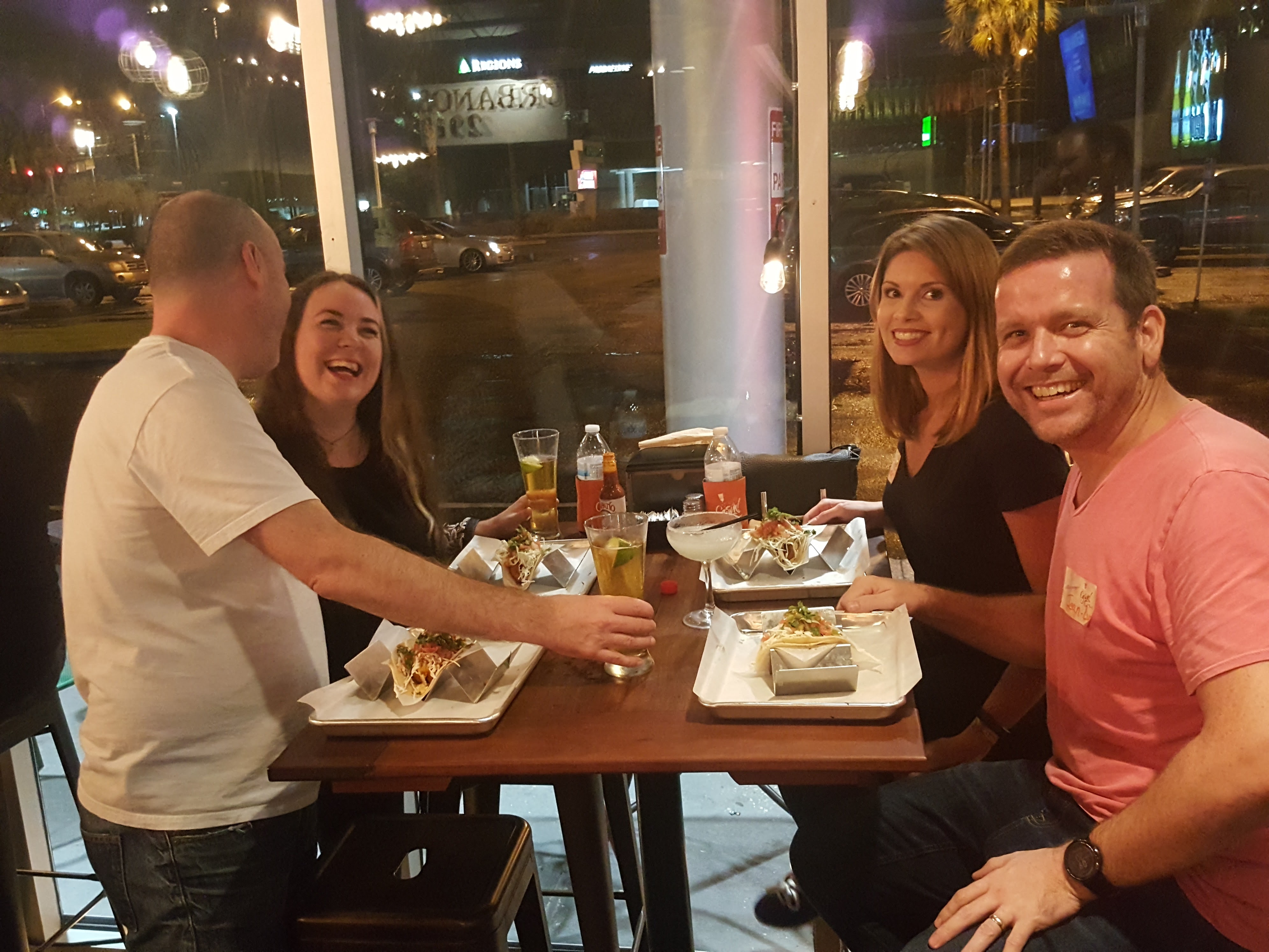 Four people smile at a table filled with trays of tacos, margaritas and beer.