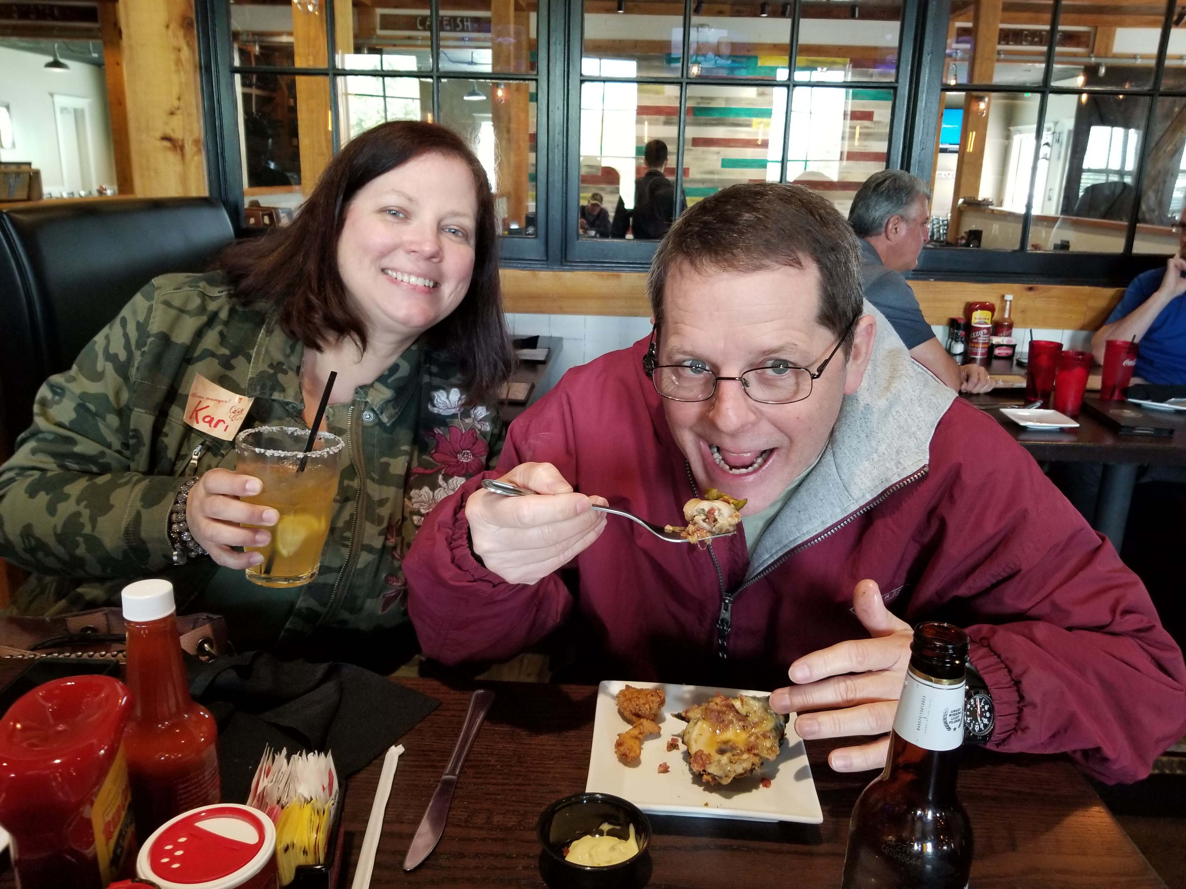 A woman smiles for the camera, holding up her salt-rimmed drink, while the man sitting next to her lifts a fork loaded with food to his mouth.