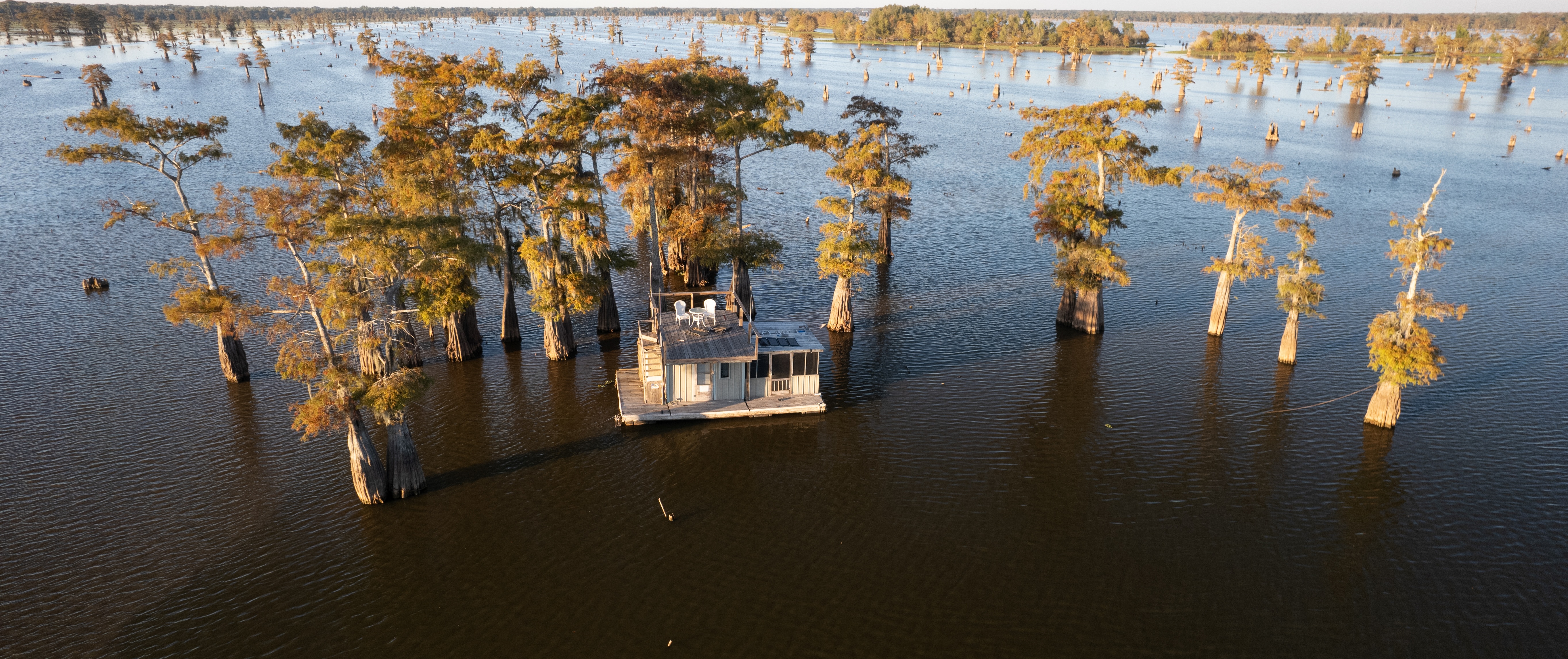 An aerial view of a wooden houseboat on the Atchafalaya Basin, anchored in a grove of massive trees.