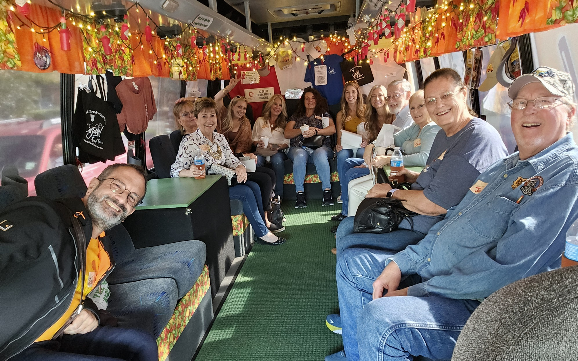 A tour group smiles for a photo on a fully decorated tour bus, with lights hanging from the ceiling and t-shirts and bags decorating the walls.