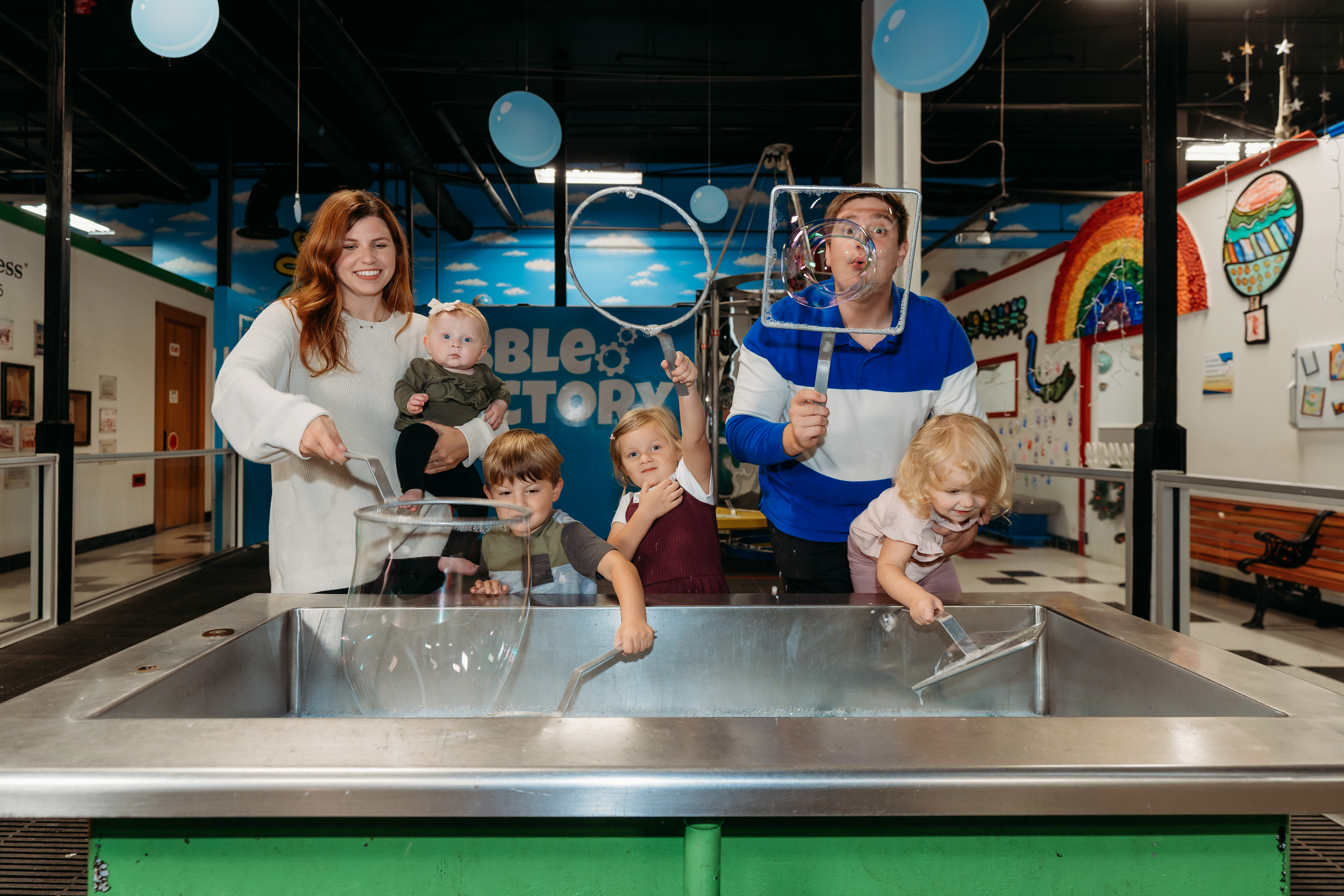 A family of six dips large metal bubble wands into a soapy solution, smiling as they wave the wands to create bubbles at the Children's Museum of Acadiana.