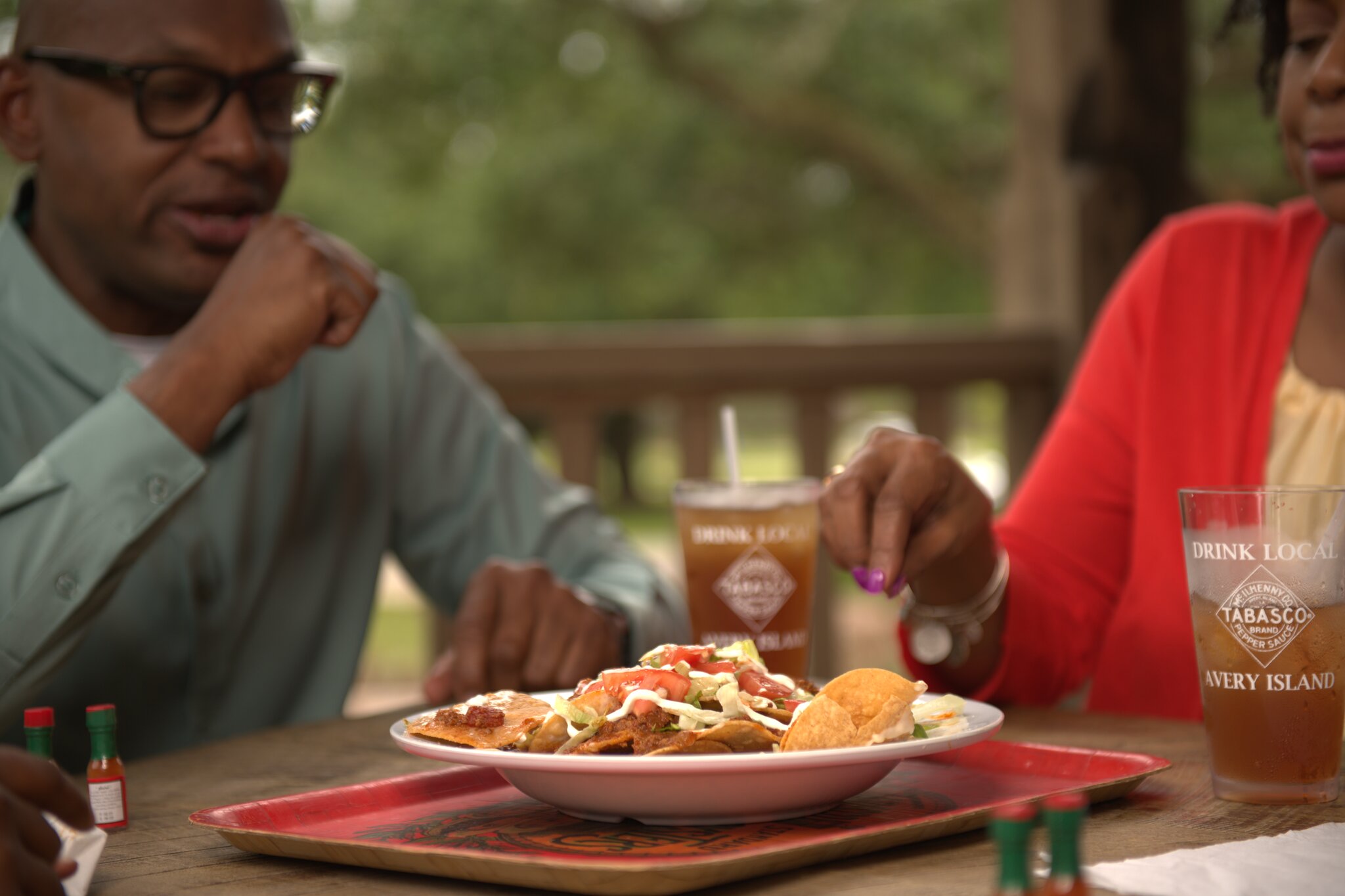 Two people share a plate of nachos at an outdoor table with iced drinks branded with "TABASCO" and "Avery Island."