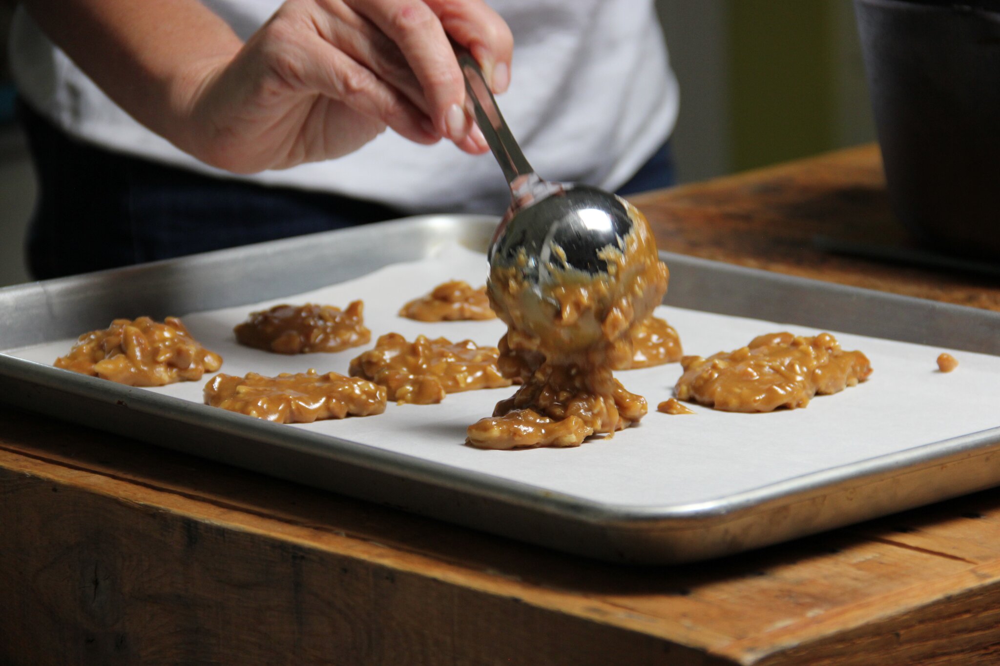 A person spoons fresh praline candy onto a baking tray lined with parchment paper.