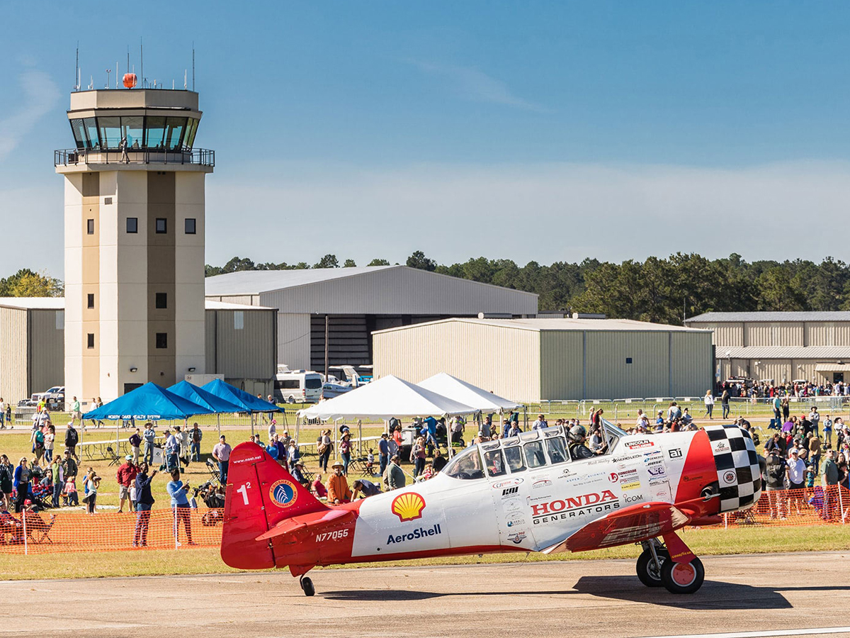 A vintage red and white AeroShell aircraft on the runway during the Hammond Northshore Regional Airshow, with crowds of spectators and the control tower in the background.