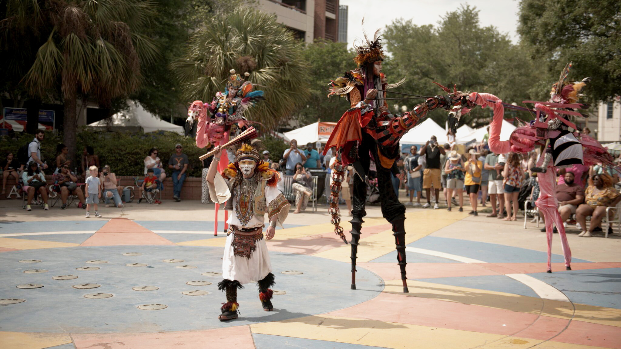 Stilt walkers and costumed performers entertain a crowd gathered in a public plaza during an outdoor festival.