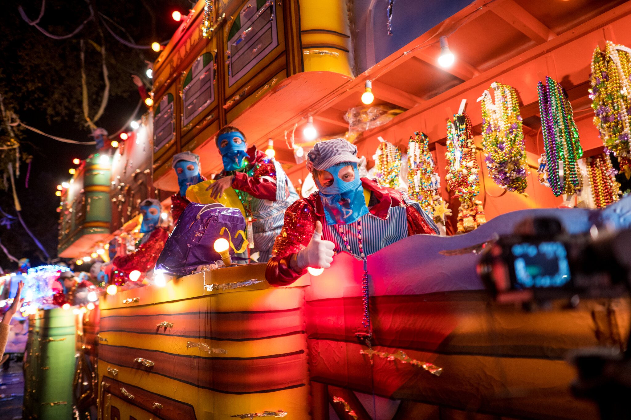 Masked Mardi Gras riders stand on a brightly lit float at night, surrounded by hanging strands of colorful beads.