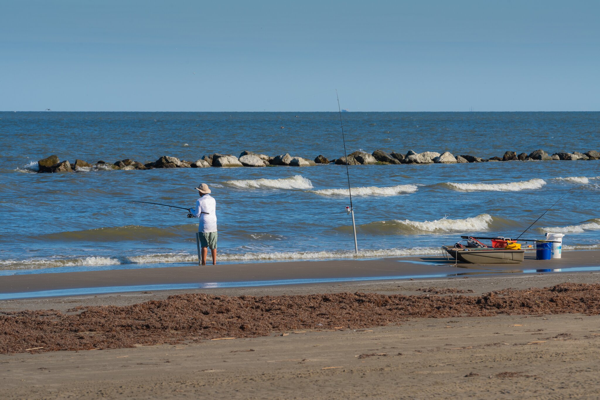 A fisherman casts a line from the beach at Grand Isle State Park with gentle waves rolling in toward a rock breakwater.