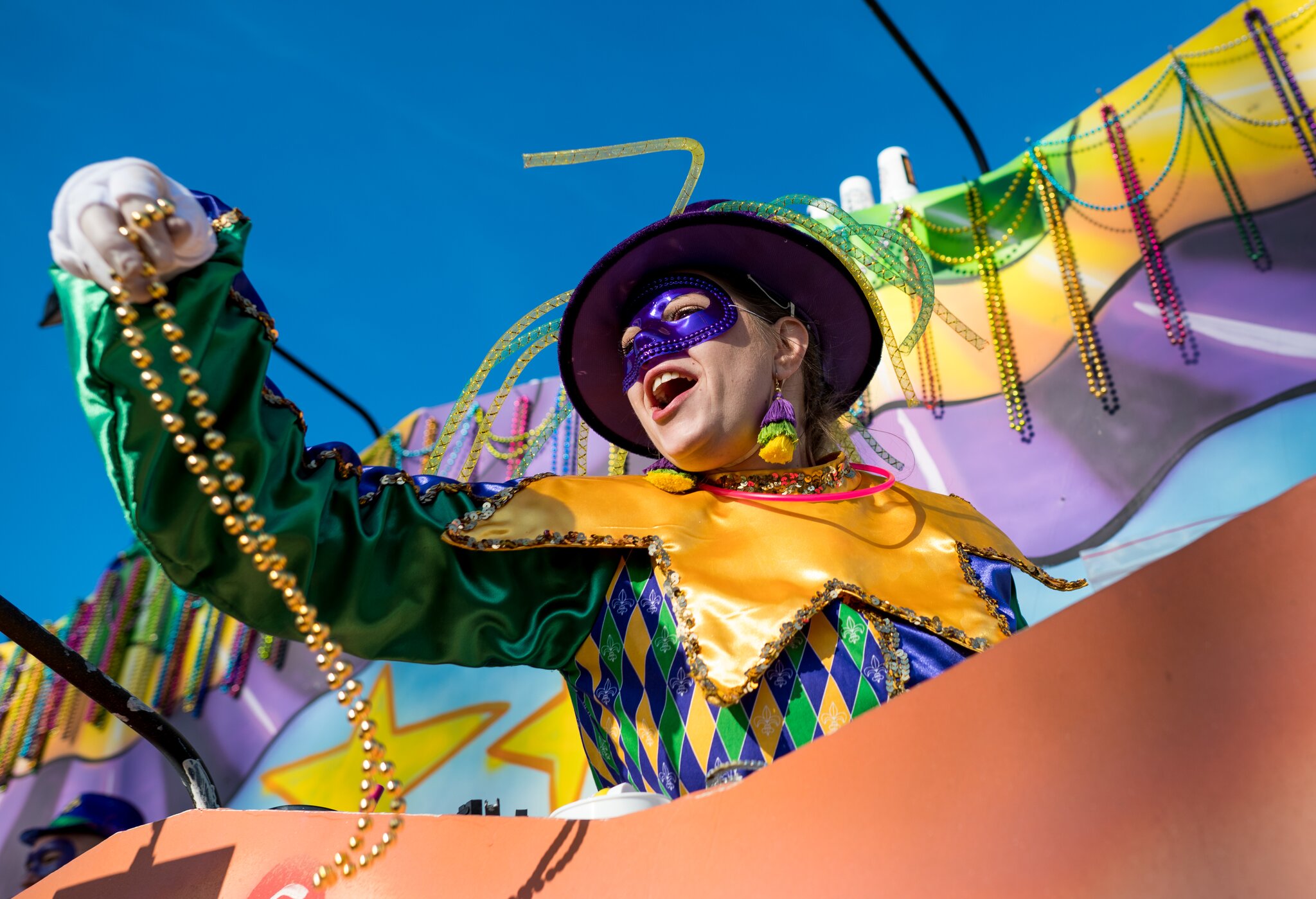 A masked parade rider tosses beads from a colorful Mardi Gras float under a clear blue sky.