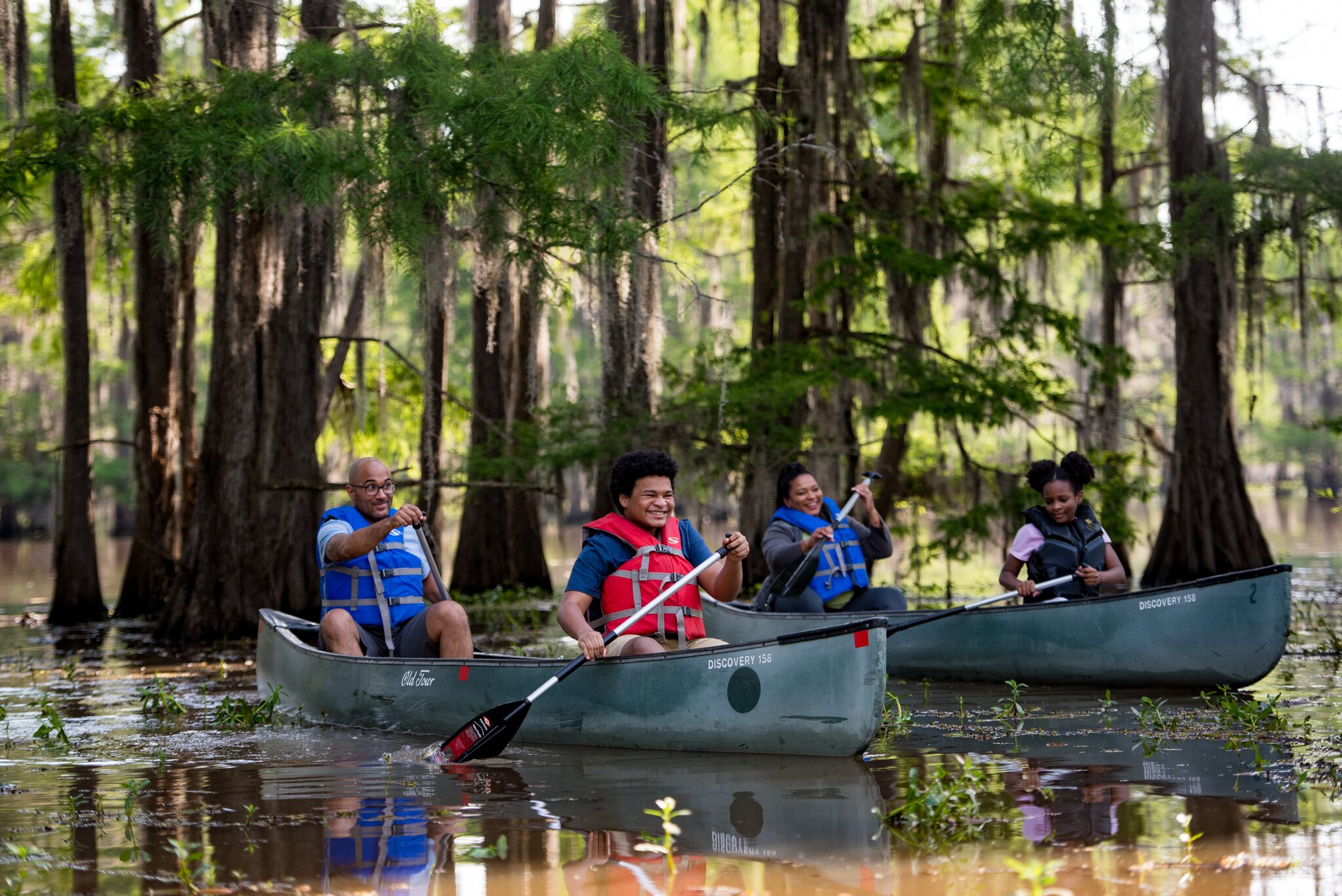 A family paddles canoes together through shallow water among cypress trees.