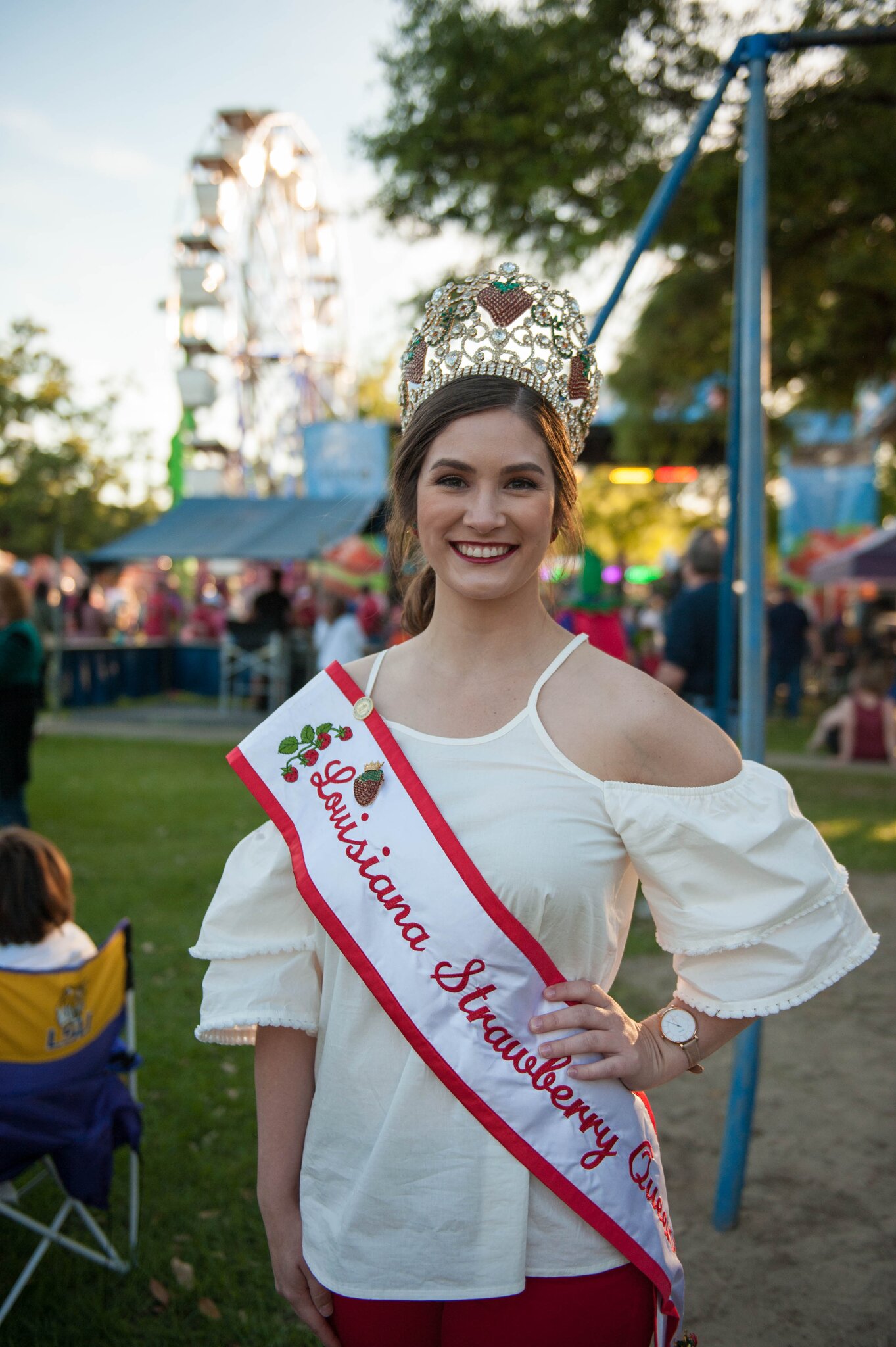 The Louisiana Strawberry Queen poses for a photo in full regalia, with a carnival in the background.