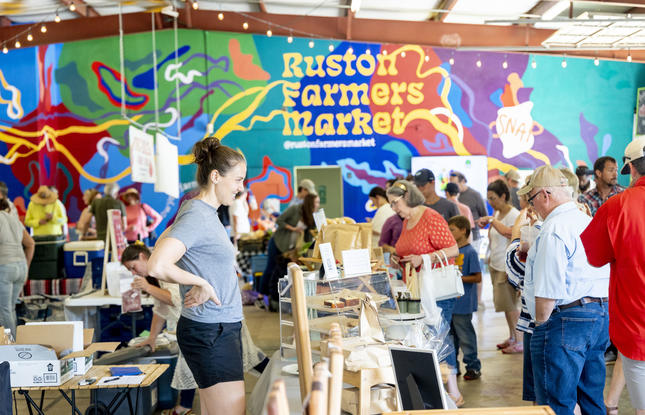 Lively crowd shopping at the busy Ruston Farmers Market in Louisiana, with a colorful mural displaying “Ruston Farmers Market” in the background.