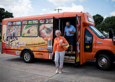 A woman in a bright orange shirt and a man in a light blue polo exit the Cajun Food Tours bus, which is painted orange with images of food printed over the windows.