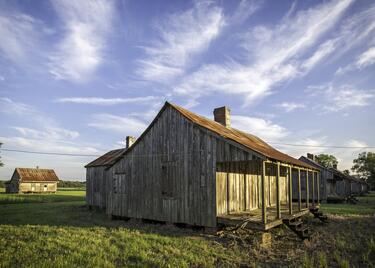 A rustic wooden cabin stands in an open field beneath a sky streaked with wispy clouds.
