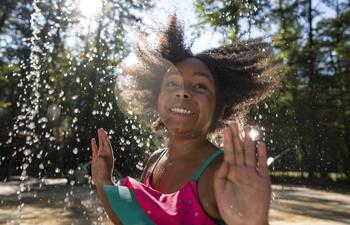 A young girl smiling while she plays in a splash pad, with water spraying around her.