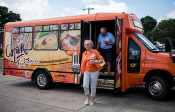 A woman in a bright orange shirt and a man in a light blue polo exit the Cajun Food Tours bus, which is painted orange with images of food printed over the windows.
