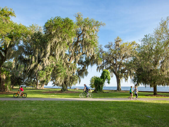 Two people ride bikes and two people jog along a paved trail on a sunny day at the Mandeville Lakefront. Credit: Louisiana Northshore