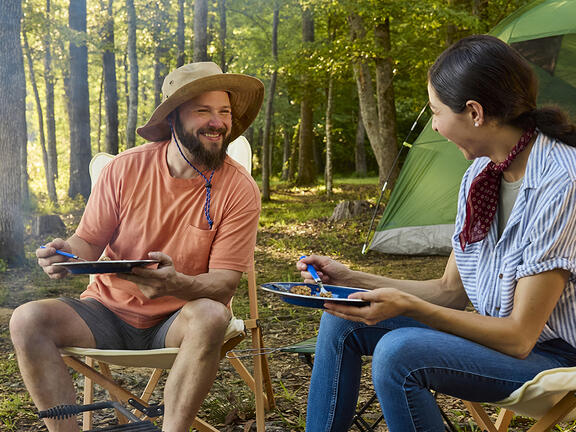 A couple sitting fire side at a camp site with tent in the background enjoying dinner at sunset
