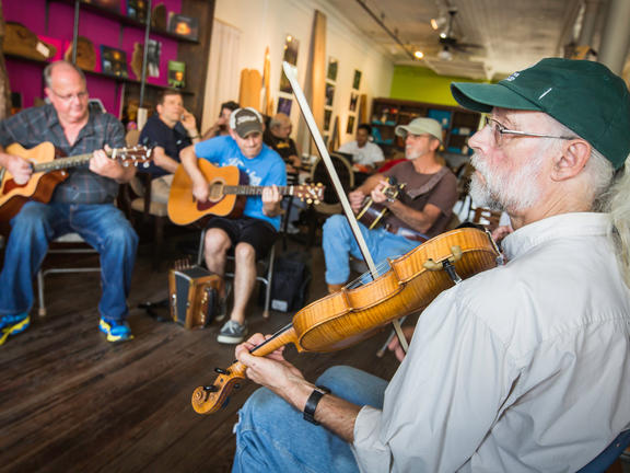 Four men sit in a circle, each holding a different instrument, including acoustic guitars and fiddles.