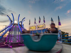 Patrons ride spinning teacups at Delcambre Shrimp Festival. Attractions glow against the soft, dusk-blue sky.