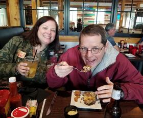 A woman smiles for the camera, holding up her salt-rimmed drink, while the man sitting next to her lifts a fork loaded with food to his mouth.
