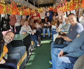 A tour group smiles for a photo on a fully decorated tour bus, with lights hanging from the ceiling and t-shirts and bags decorating the walls.