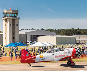 A vintage red and white AeroShell aircraft on the runway during the Hammond Northshore Regional Airshow, with crowds of spectators and the control tower in the background.
