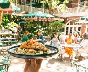 A hand holds a plated seafood dish above an outdoor courtyard at Brennan’s in New Orleans, with green café tables, striped umbrellas, and diners seated in the background.