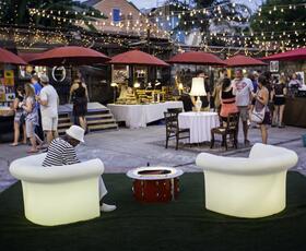 A lively nighttime scene on Frenchmen Street shows people browsing art vendors and socializing under string lights and red umbrellas.