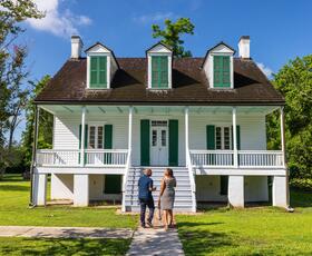 A couple walks toward a classic white plantation-style home with green shutters under a bright blue sky.
