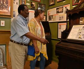Two visitors explore a museum room filled with framed photographs, historical text panels and a vintage upright piano.