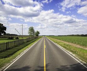 A long, straight rural road cuts through flat farmland under a wide sky filled with scattered clouds.