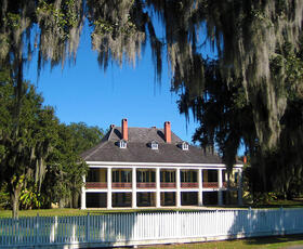 Front view of the Destrehan Plantation main house with white columns and moss-draped trees, surrounded by a white wooden fence.
