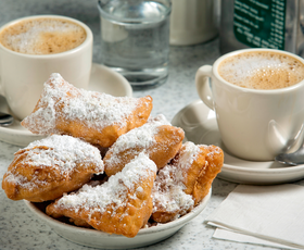Two white mugs holding cafe au lait are set beside a shallow bowl of flaky beignets topped with powdered sugar from Cafe du Monde in New Orleans, Louisiana.