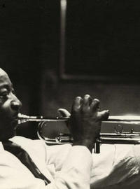 A side view of Dave Bartholomew blowing big cheeked into a trumpet in a dark room. 