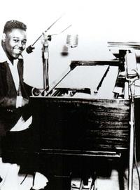 Fats Domino smiles at the camera from his piano, where he sits in front of a microphone